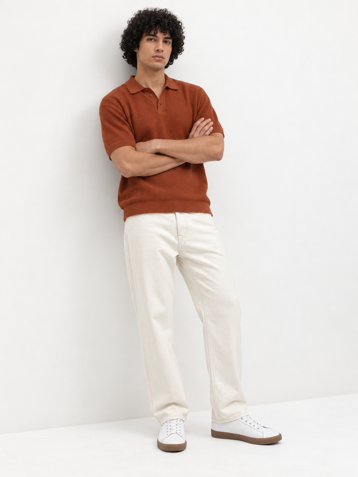 A man with curly dark hair stands against a white wall wearing a rust-orange short-sleeve polo shirt, off-white trousers, and white sneakers with brown soles.
