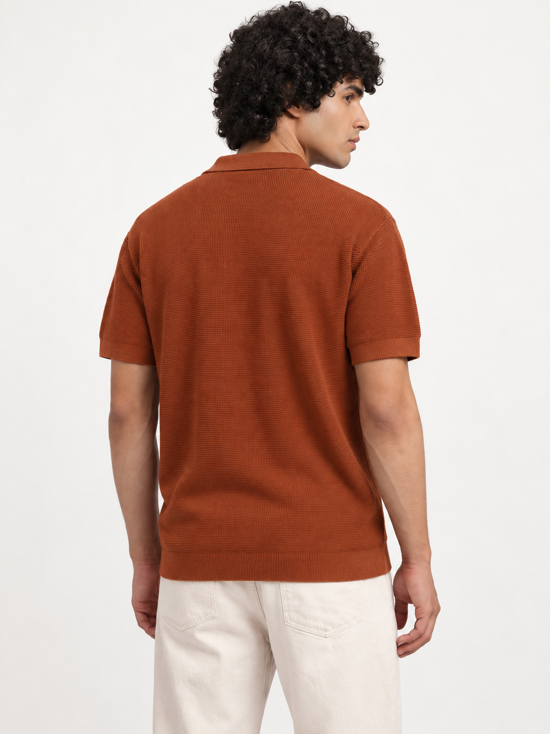 A man with curly dark hair is shown from the back wearing a rust-orange short-sleeve knit polo shirt and off-white trousers, photographed against a clean white studio background.
