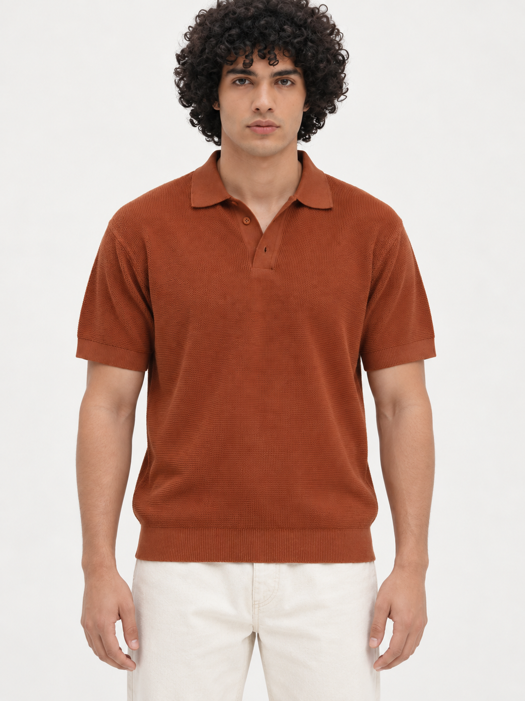 A man with curly dark hair wearing a rust-orange short-sleeve knit polo shirt and off-white trousers, standing against a plain light gray studio background.