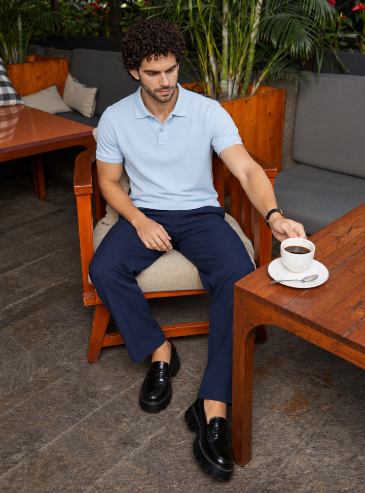 A man sitting in a wooden lounge chair at an outdoor cafe, wearing a light blue polo shirt, navy trousers, and black loafers, reaching for a cup of coffee on a wooden table.