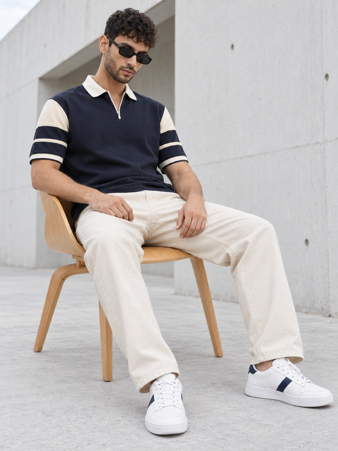 A man wearing a navy and beige short-sleeve polo t-shirt, cream trousers, and white sneakers sits on a wooden chair against a concrete wall.