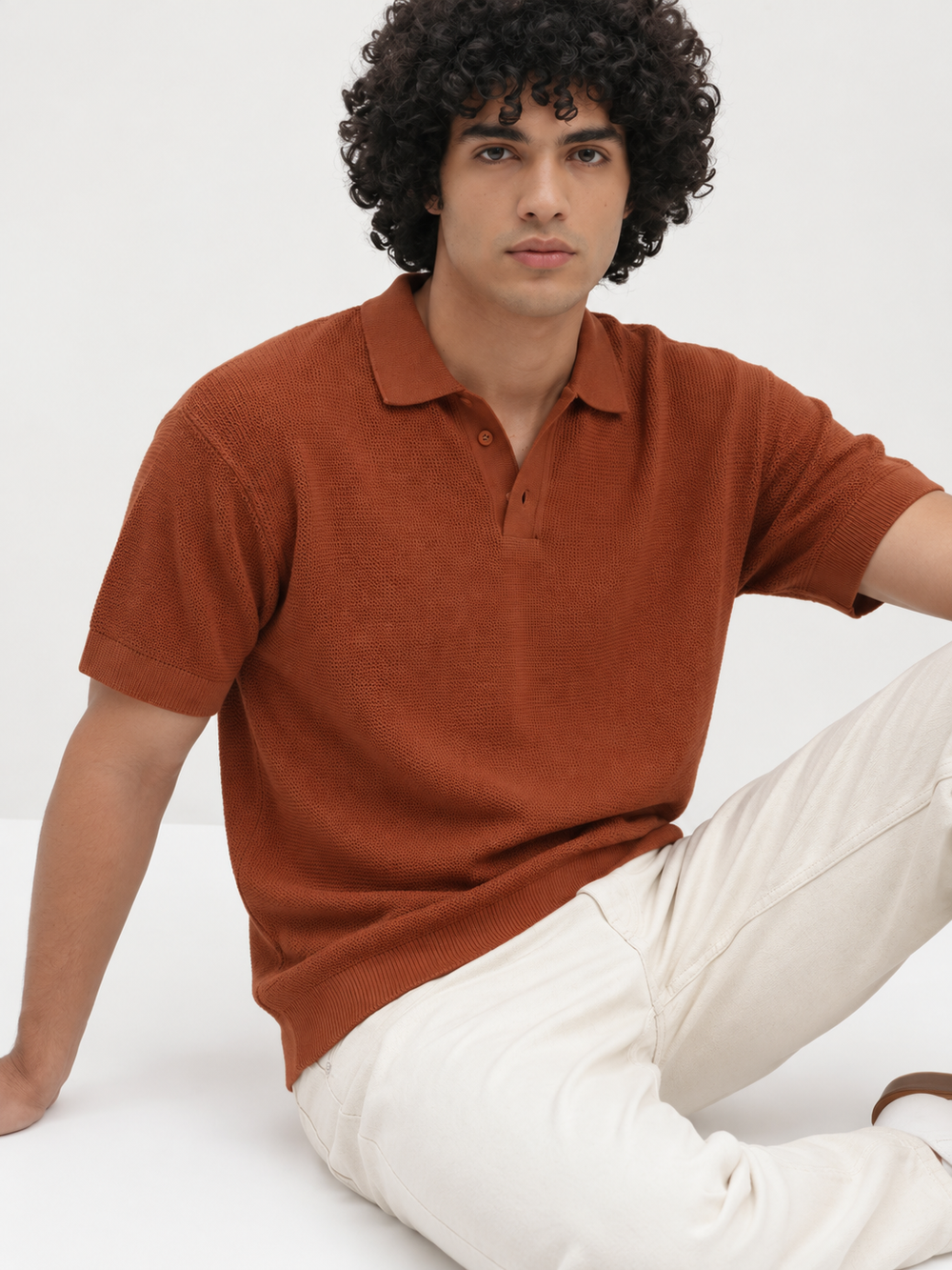 A man with curly black hair wearing a rust-orange short-sleeve knit polo shirt and off-white trousers, sitting against a plain white studio background.