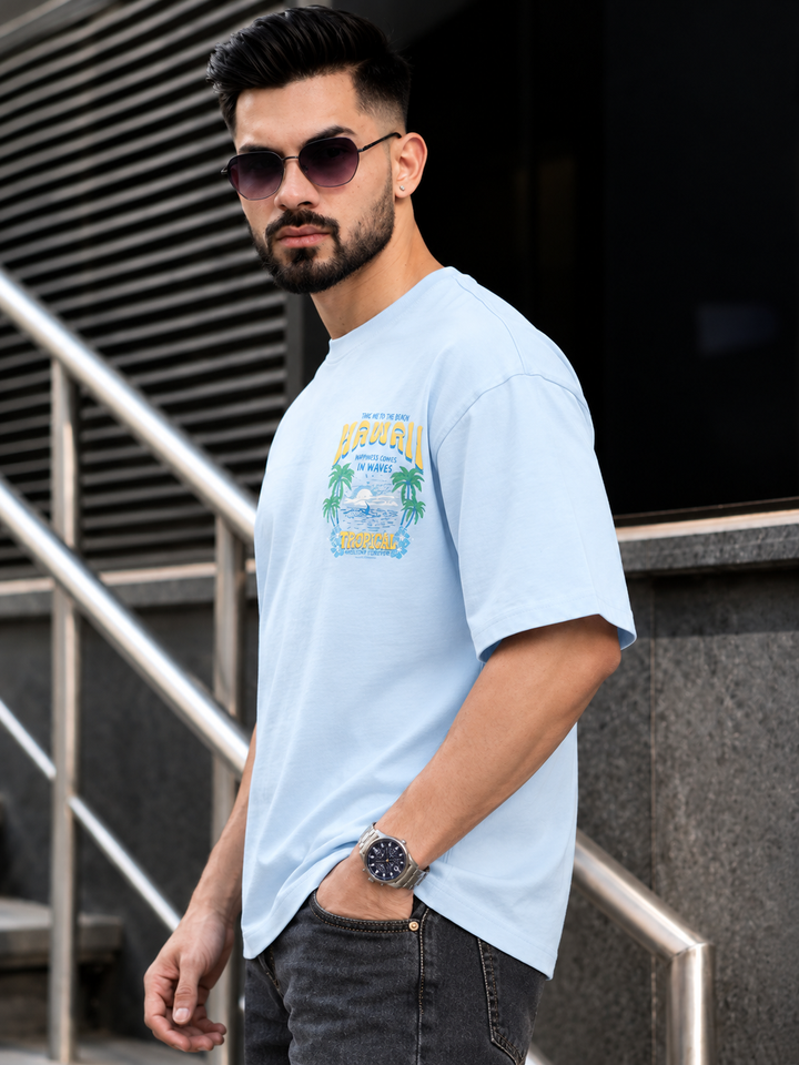 A man wearing a light blue graphic t-shirt with a tropical beach design and the word 'HARAVI' printed on the chest, paired with dark gray jeans and sunglasses, standing in front of a modern building staircase.
