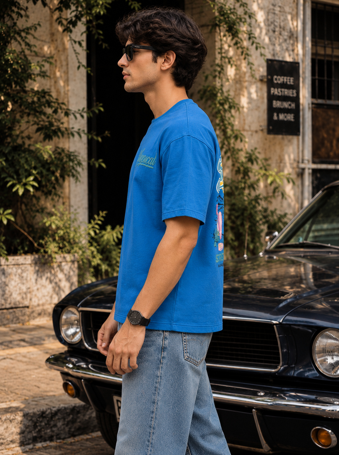 A man wearing a bright blue graphic t-shirt and light wash jeans stands in profile next to a black vintage car in front of a rustic building with greenery.