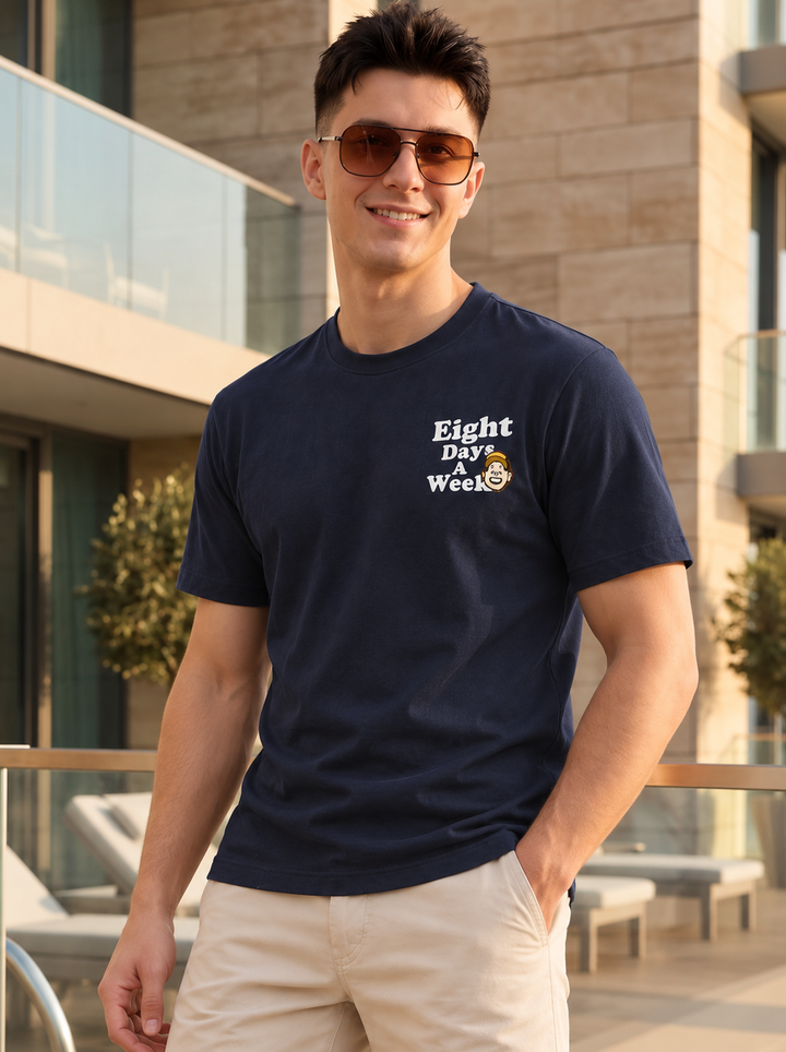 A man wearing a navy blue crewneck t-shirt with a white graphic that reads 'Eight Day Week' and a small cartoon character, paired with beige shorts, standing outdoors in front of a modern building.