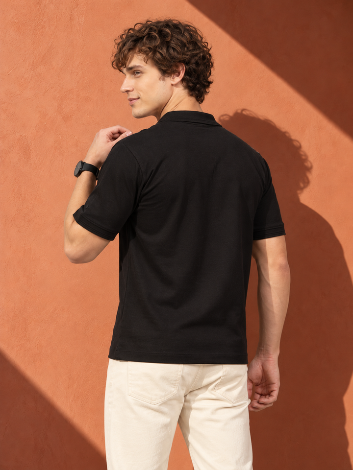 A man wearing a black short-sleeve polo shirt and off-white trousers, photographed from the back against a textured terracotta wall with strong diagonal shadows.