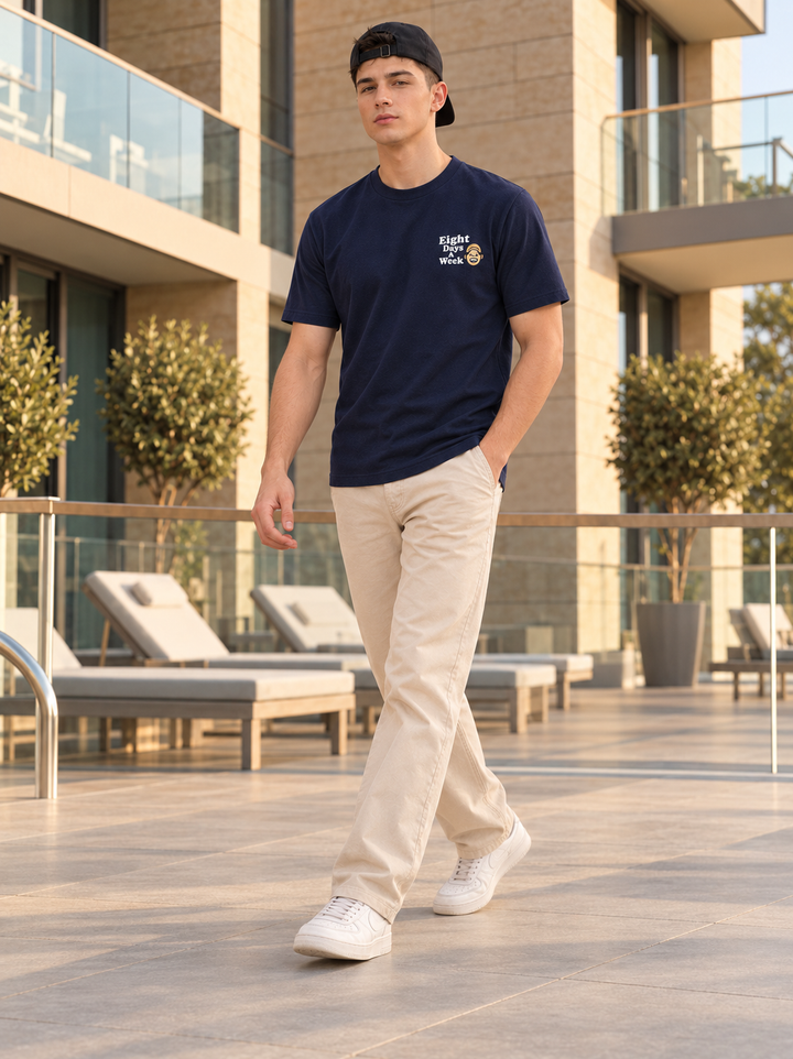 A man walking on a modern outdoor terrace wearing a navy blue t-shirt with a small white graphic on the left chest, beige trousers, and white sneakers.