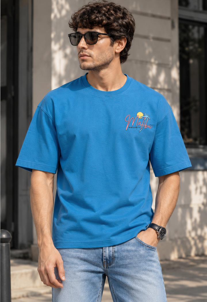A man wearing a bright blue crewneck t-shirt with a small red and yellow graphic on the left chest, paired with light-wash blue jeans, standing outdoors in front of a building.