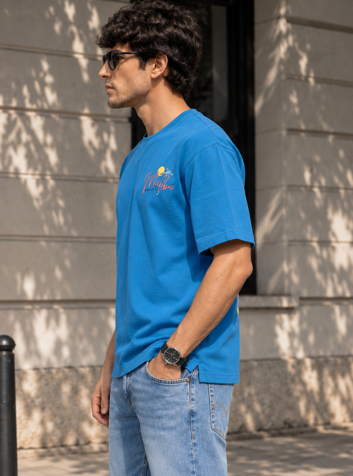 A man wearing a bright blue crewneck t-shirt with a small graphic on the left chest, paired with light blue jeans, standing outdoors against a stone wall.