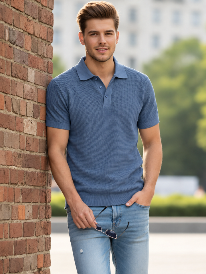 A man wearing a blue short-sleeve polo shirt and light blue jeans stands outdoors next to a red brick wall, holding sunglasses.