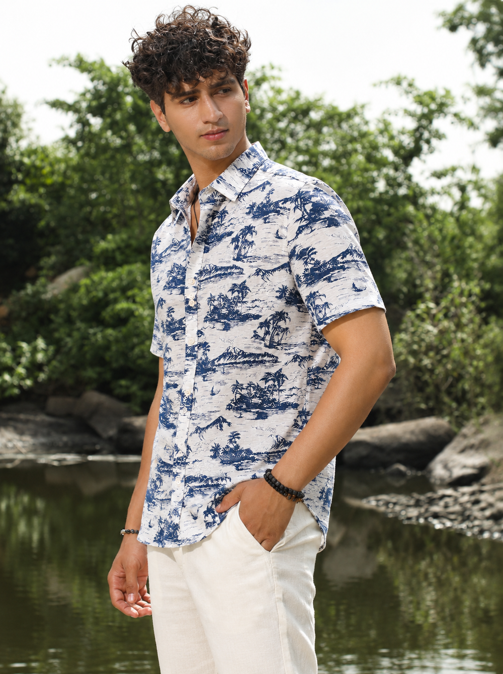 A man wearing a blue and white tropical patterned short-sleeve button-down shirt and off-white pants, standing outdoors near a body of water with trees in the background.