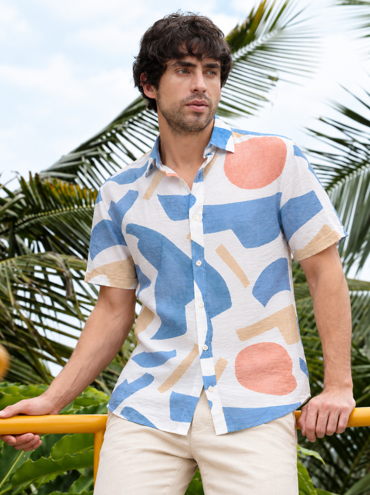 A man wearing a short-sleeve button-up shirt with an abstract geometric pattern in blue, beige, and coral pink on a white base, paired with light beige pants, set against a tropical background with palm leaves.