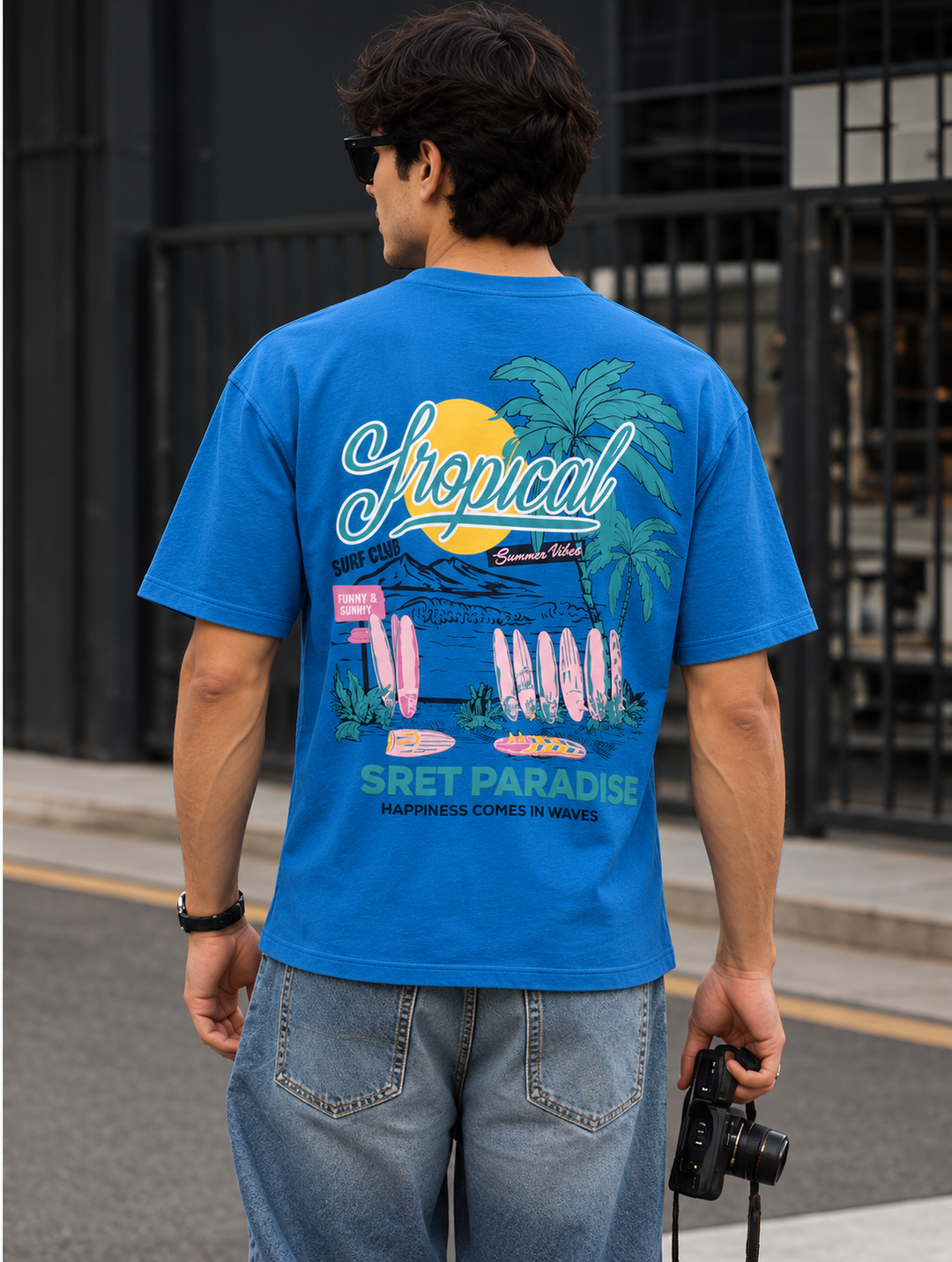 A man wearing a bright blue graphic mentor t-shirt with a tropical beach print on the back, paired with light wash jeans, standing outdoors on a street.