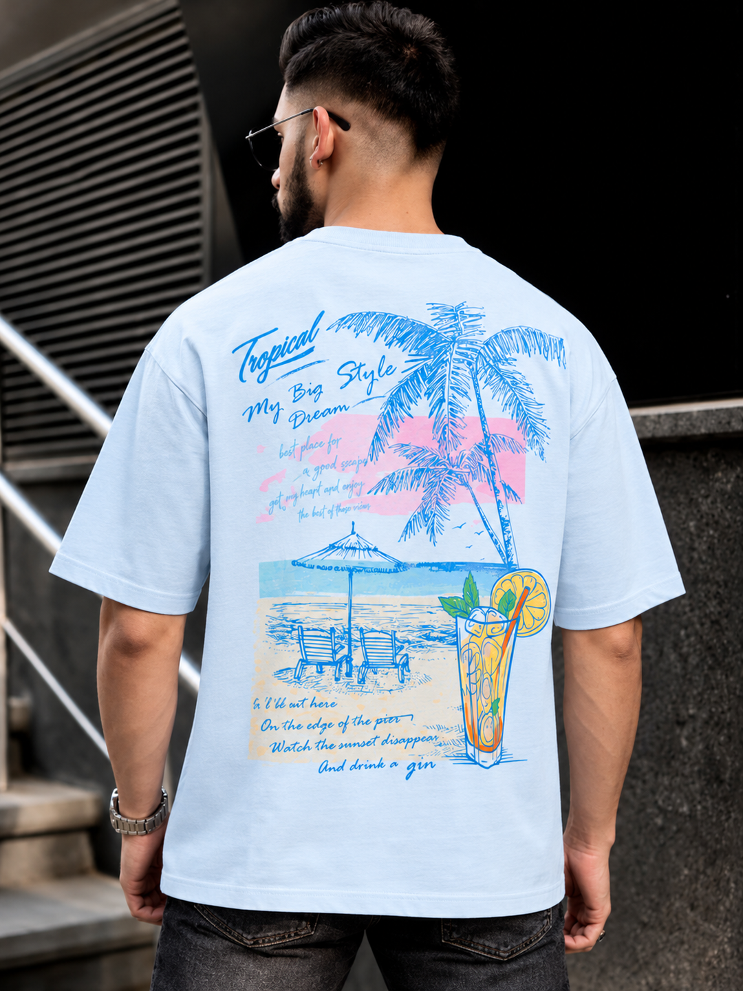 A man viewed from behind wearing a light blue graphic t-shirt with a tropical beach design and text, paired with dark jeans, standing outdoors against a dark textured wall.
