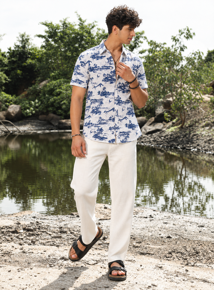 A man wearing a blue and white tropical print short-sleeve button-down shirt, white linen pants, and black sandals, standing on a rocky shore next to a calm lake with trees in the background.