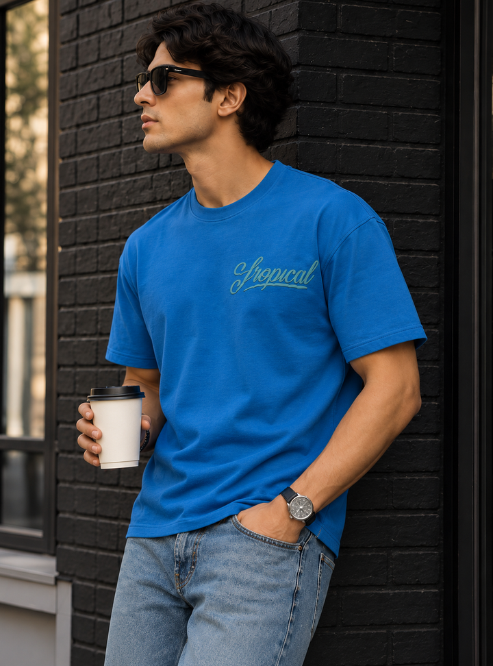 A man wearing a bright blue crewneck t-shirt by mentor with a light blue 'Tropical' script logo on the chest, paired with light wash jeans and sunglasses, holding a coffee cup against a black brick wall.