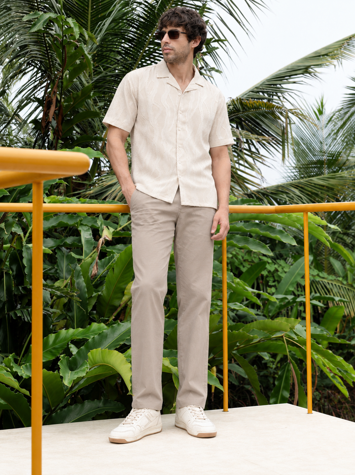 A man wearing a light beige short-sleeve button-up shirt, beige trousers, and white sneakers stands on a platform with a yellow railing, surrounded by lush tropical green foliage.